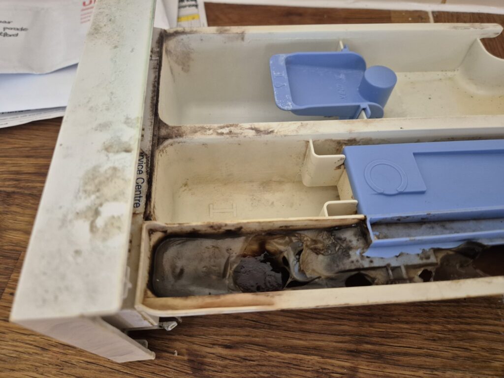 Heavily soiled washing machine detergent drawer with built-up residue and dark grime before cleaning